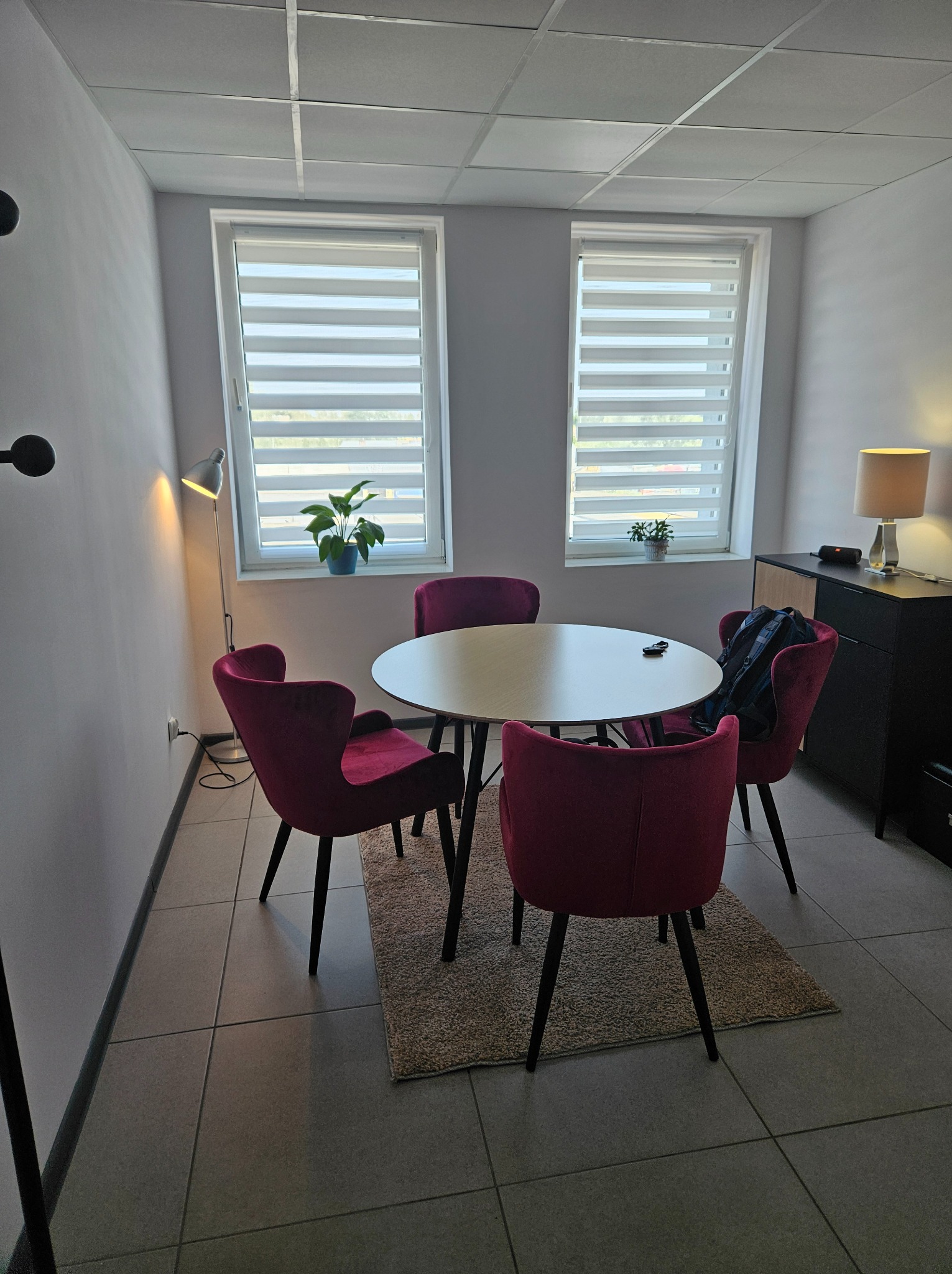 Jasne pomieszczenie z okrągłym stołem, czterema bordowymi krzesłami, lampą podłogową i komodą. Dwa okna z żaluzjami w tle.