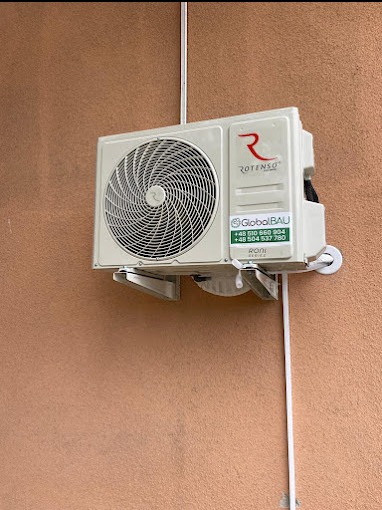 Biała jednostka zewnętrzna klimatyzatora Rotenso zamontowana na beżowej ścianie budynku, z widocznymi rurami i mocowaniami, z naklejką z numerami telefonów serwisu GlobalBAU.