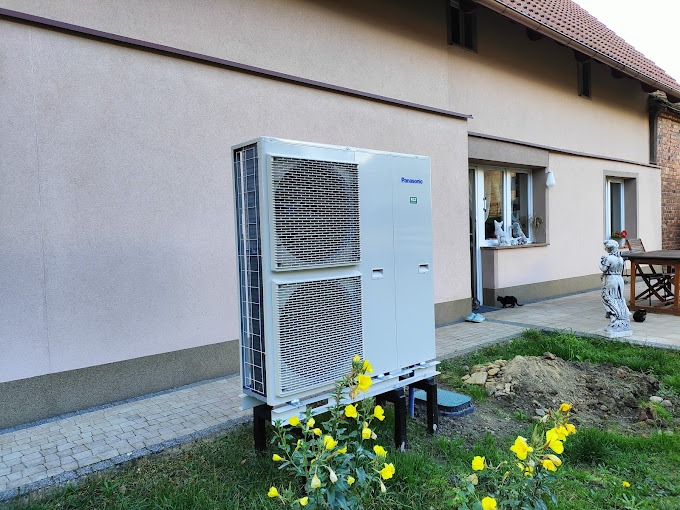 Biała pompa ciepła Panasonic zamontowana na drewnianych wspornikach przed jasnym budynkiem z oknem, na trawniku kwitną żółte kwiaty.