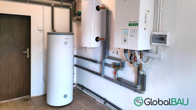 Białe zbiorniki i urządzenie grzewcze zamontowane na ścianie w pomieszczeniu technicznym, z widocznymi rurami izolowanymi termicznie i skrzynką elektryczną.