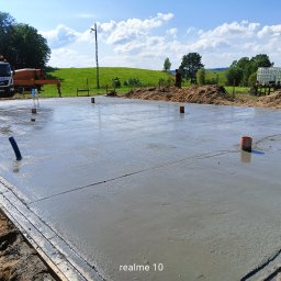 GoodJob - Świeżo wylany fundament z widocznymi elementami instalacji i ciężarówką do betonu w tle, na tle zielonego krajobrazu pod błękitnym niebem.