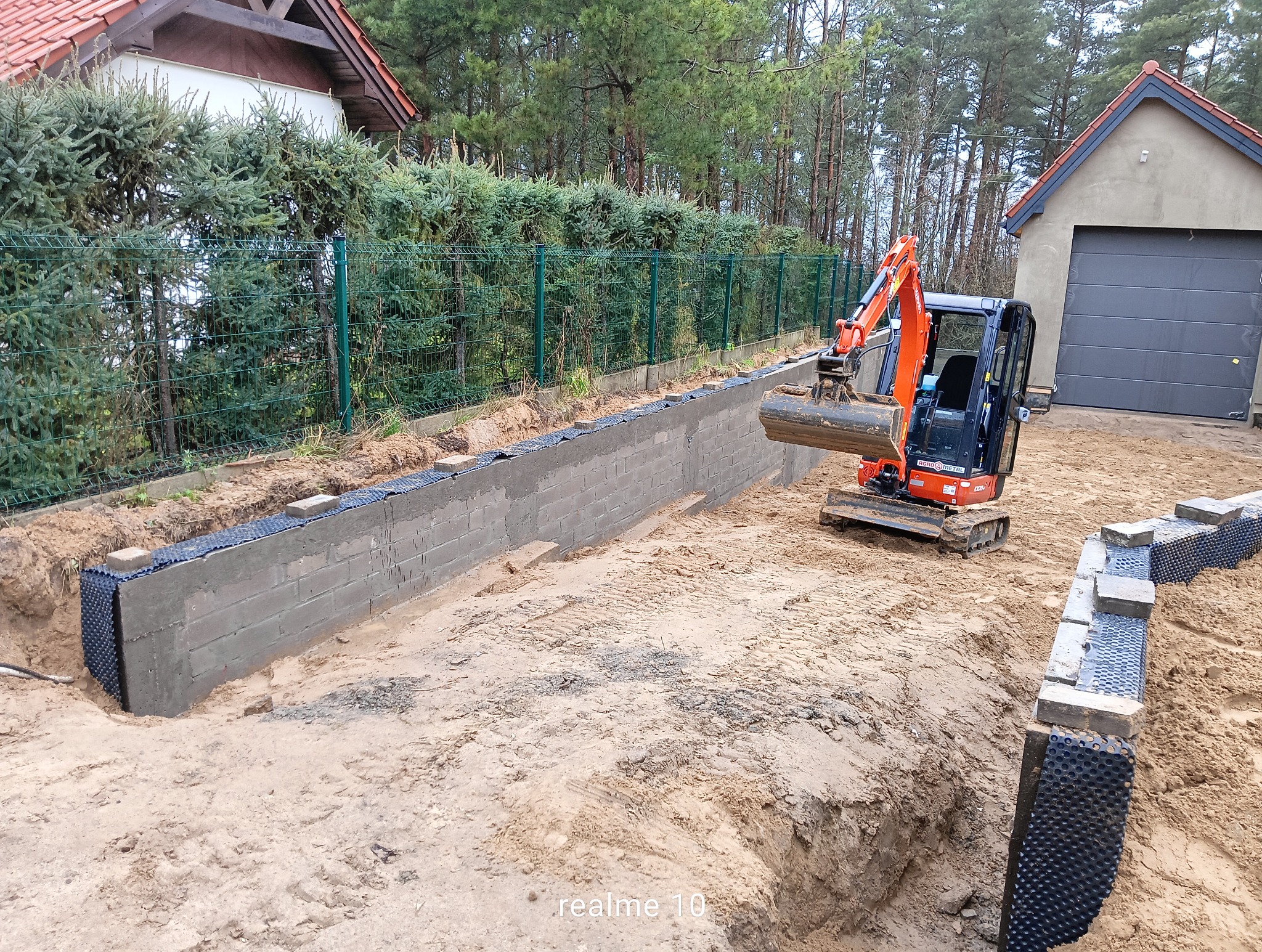 Wykop pod fundamenty z częściowo wzniesioną ścianą z bloczków betonowych, zabezpieczoną folią kubełkową. Mała koparka na placu budowy. Widok na posesję z zielonym ogrodzeniem.