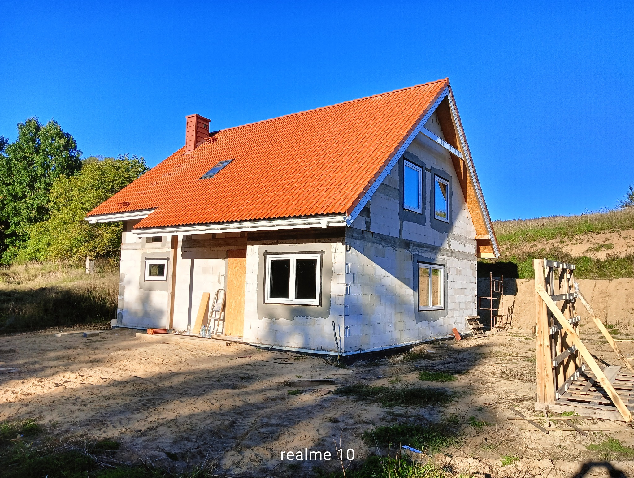 Dom w stanie surowym otwartym z czerwoną dachówką, widoczne okna i elementy konstrukcyjne. Na pierwszym planie drewniane szalunki.