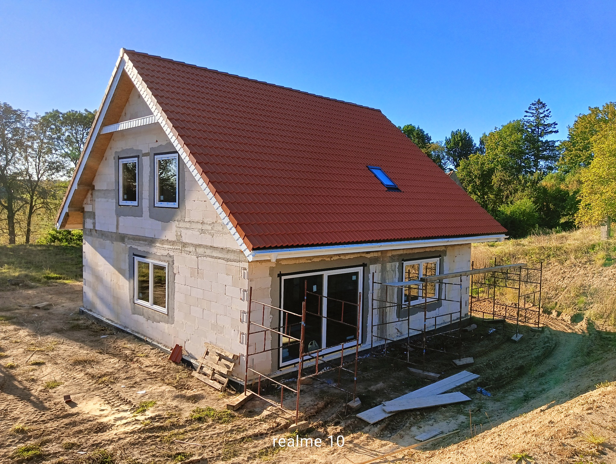 Surowy dom jednorodzinny z czerwonym dachem, widoczne rusztowania i teren budowy. Okna wstawione, ściany z białego bloczka. Błękitne niebo w tle.