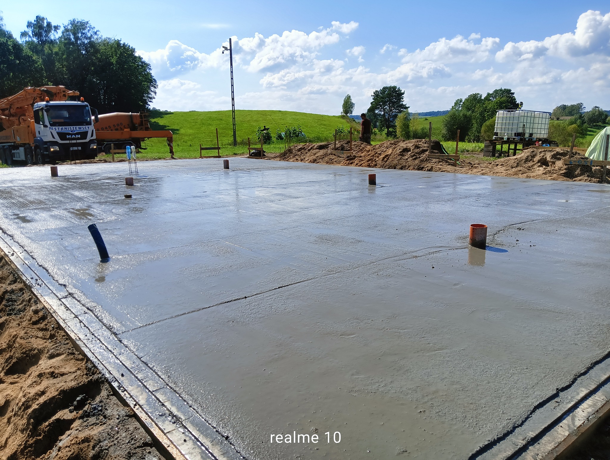 Świeżo wylany fundament z widocznymi elementami instalacji i ciężarówką do betonu w tle, na tle zielonego krajobrazu pod błękitnym niebem.