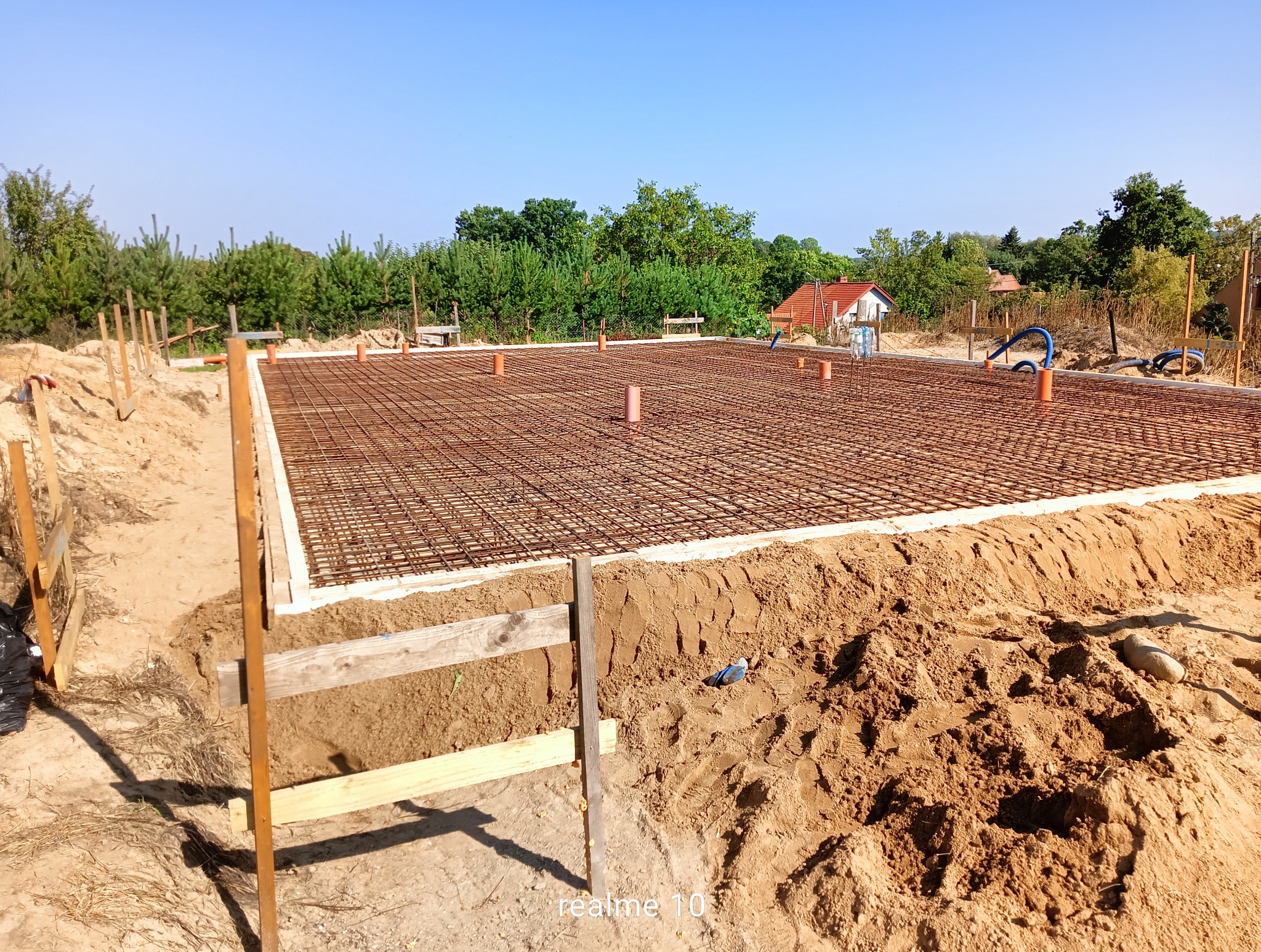 Zbrojenie fundamentów na wykopie, widoczne szalunki i wystające rury. W tle zieleń i zabudowania. Słoneczny dzień.
