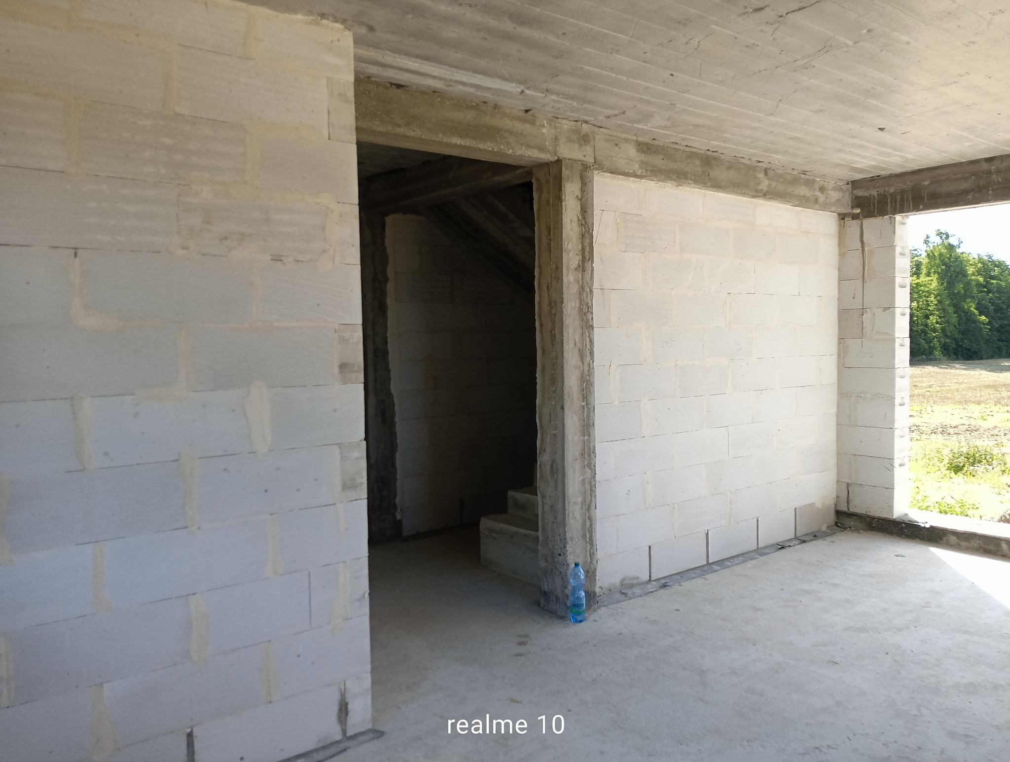 Surowy stan budynku w trakcie budowy: ściany z białych bloczków, betonowy strop, otwór drzwiowy prowadzący do schodów. Butelka wody przy ścianie.