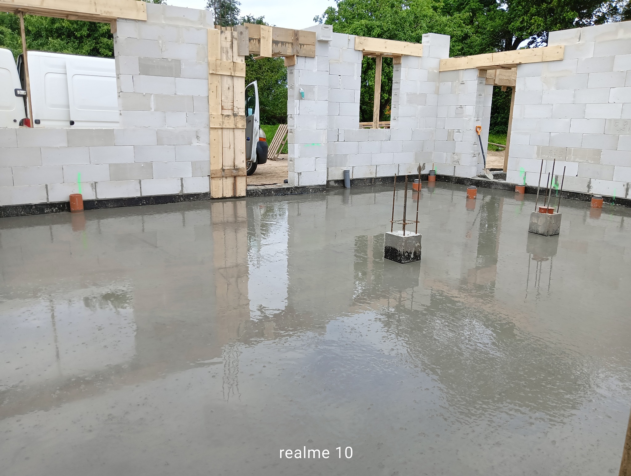 Wylany fundament z wystającymi prętami zbrojeniowymi, ściany z bloczków betonowych w trakcie budowy. Widoczny samochód dostawczy w tle.