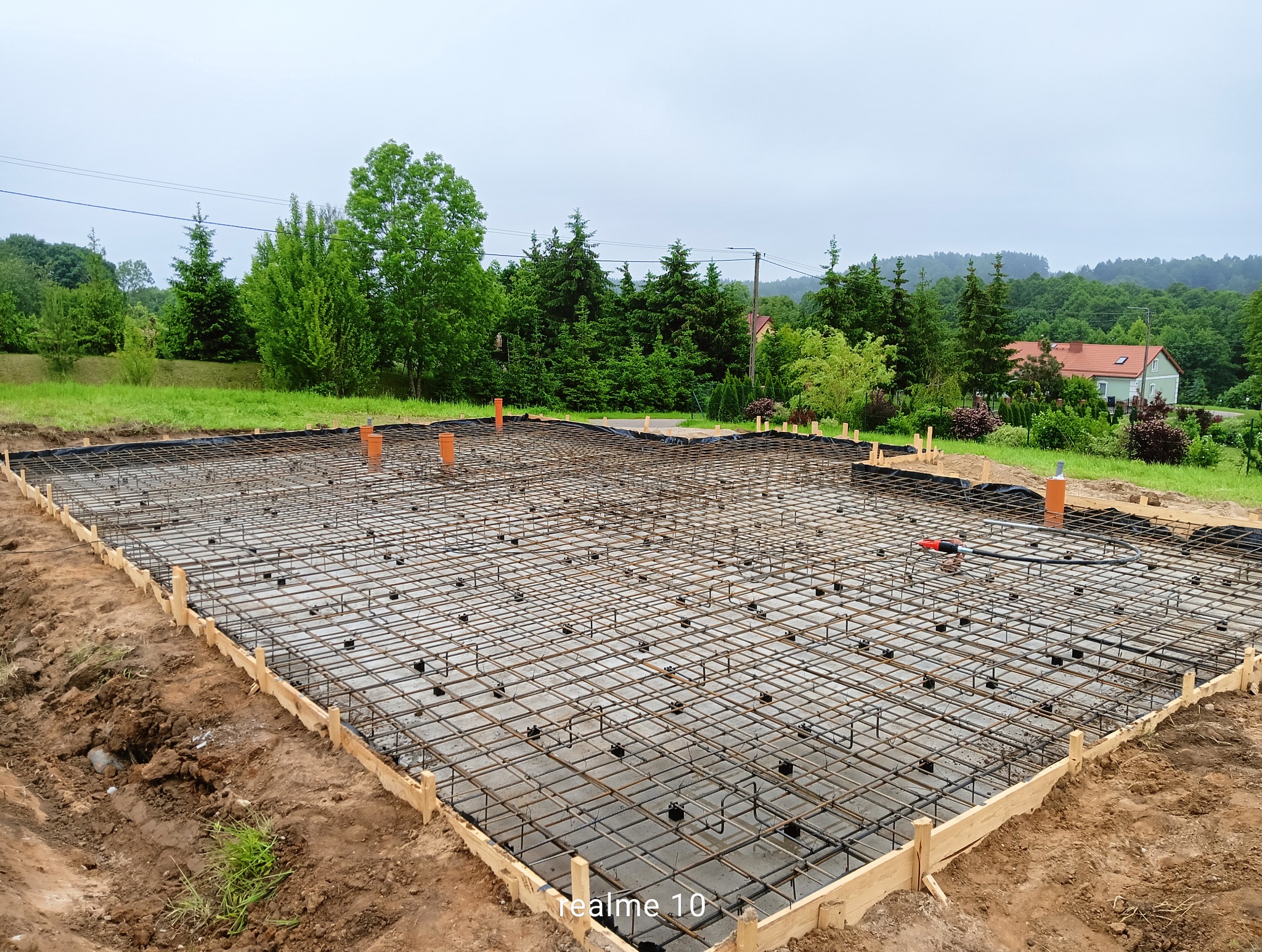 Zbrojenie fundamentu domu jednorodzinnego z widocznymi rurami instalacyjnymi i drewnianym szalunkiem, na tle zieleni i zabudowań. Widok z góry, pochmurny dzień.