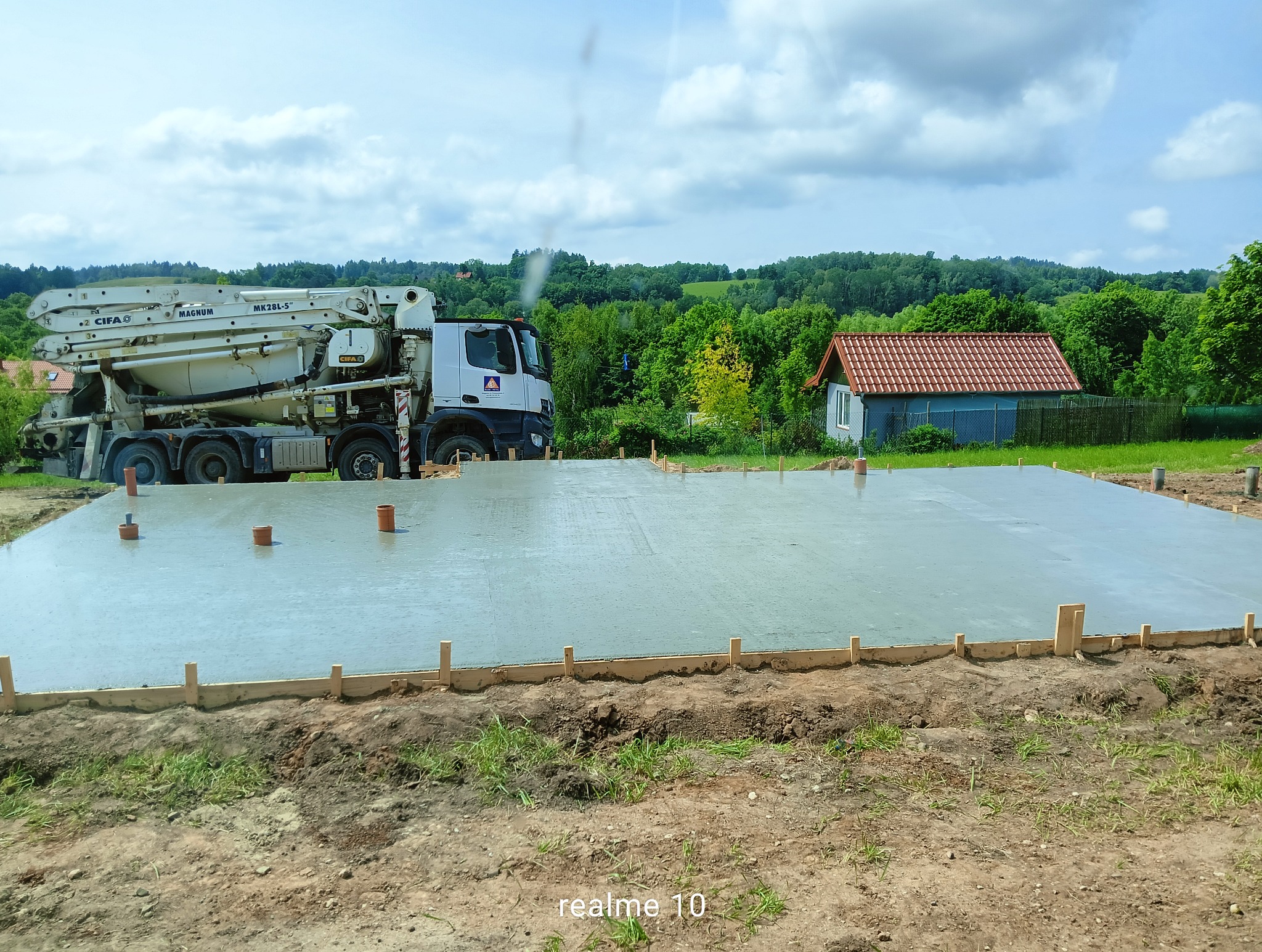Świeżo wylany fundament pod budynek z widocznym szalunkiem i rurami wentylacyjnymi, obok stoi betoniarka. W tle zielony krajobraz i mały budynek.