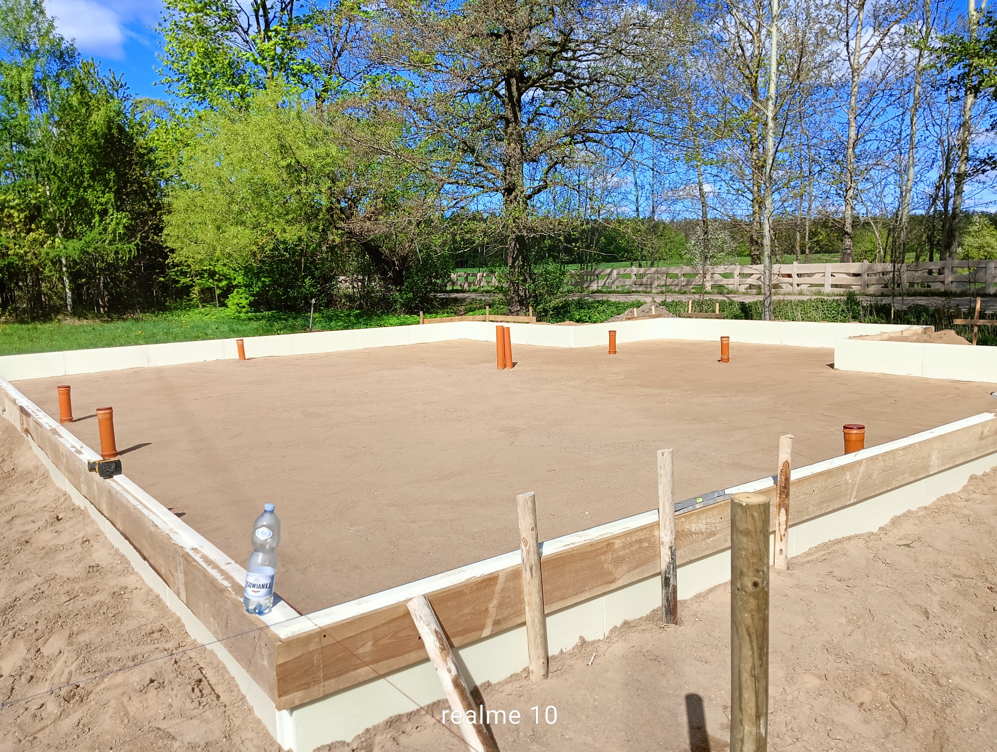 Przygotowany teren pod fundamenty z izolacją, widoczne rury kanalizacyjne i butelka wody. Tło: drzewa i ogrodzenie. Ujęcie z poziomu gruntu.