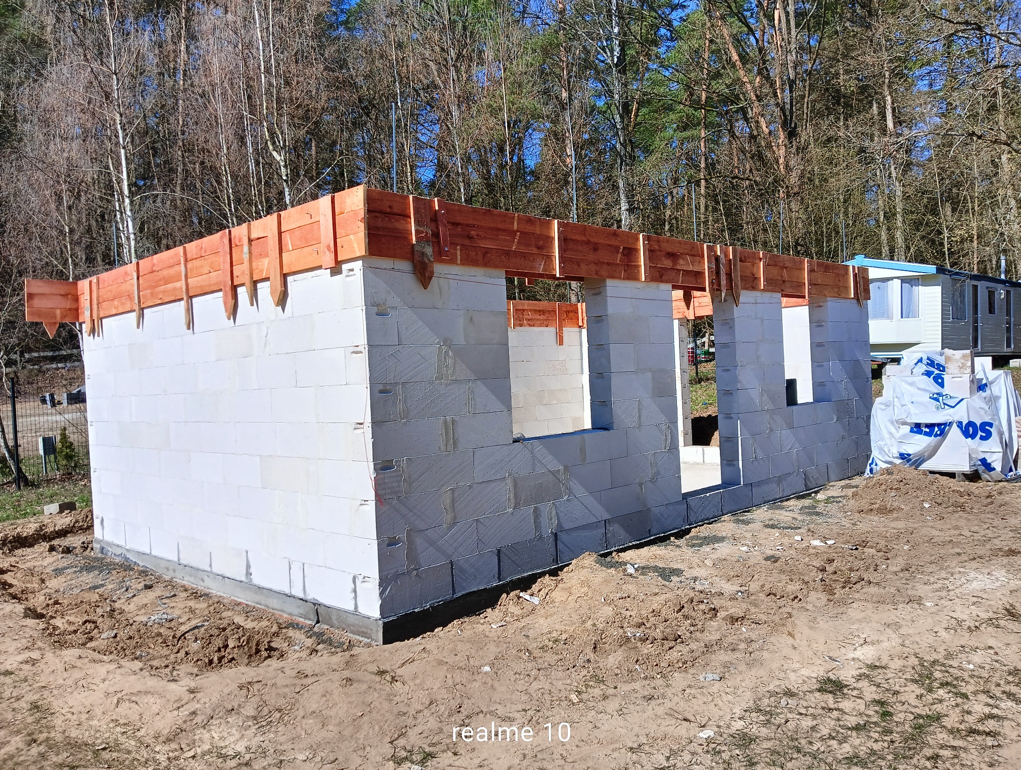 Wznoszenie ścian parteru z bloczków betonu komórkowego Ytong, z widocznym szalunkiem drewnianym na górze, przygotowanym pod wylanie wieńca. W tle drzewa i przyczepa kempingowa.