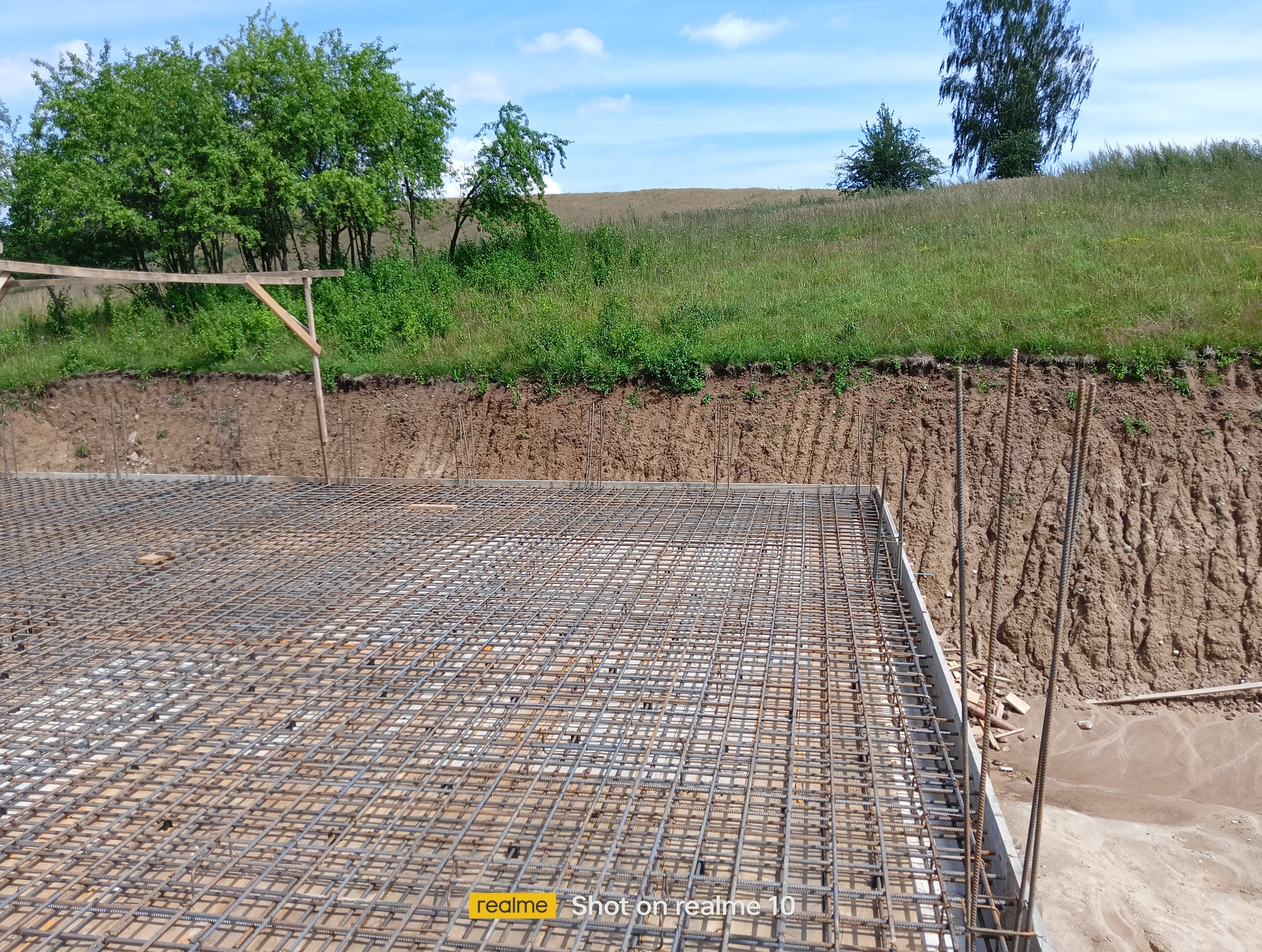 Zbrojenie fundamentów w Mrągowie, widok z góry na siatkę stalową i wystające pręty, przygotowane do zalania betonem. W tle ziemia i zieleń.