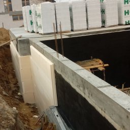 MONTOWNIA DOMÓW SPÓŁKA Z OGRANICZONĄ ODPOWIEDZIALNOŚCIĄ - Budowa fundamentów: ściana fundamentowa z izolacją termiczną, zabezpieczona hydroizolacją, z widocznymi zbrojeniami i paletami bloczków betonowych H+H na górze.