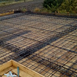 MONTOWNIA DOMÓW SPÓŁKA Z OGRANICZONĄ ODPOWIEDZIALNOŚCIĄ - Zbrojenie płyty fundamentowej z deskowaniem i narzędziami w rogu, w tle budowa domu jednorodzinnego w stanie surowym otwartym.