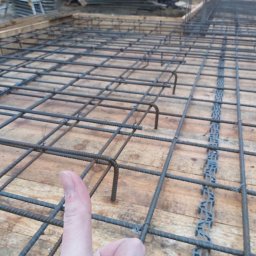 MONTOWNIA DOMÓW SPÓŁKA Z OGRANICZONĄ ODPOWIEDZIALNOŚCIĄ - Zbrojenie fundamentu ułożone na deskowaniu z widocznym elementem dystansowym i kciukiem dającym perspektywę skali.