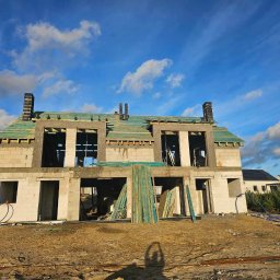 MONTOWNIA DOMÓW SPÓŁKA Z OGRANICZONĄ ODPOWIEDZIALNOŚCIĄ - Dwukondygnacyjny budynek w stanie surowym otwartym z widocznym drewnianym szkieletem dachu, kominami z cegły klinkierowej i materiałami budowlanymi na placu budowy w słoneczny dzień.