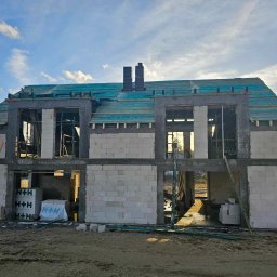 MONTOWNIA DOMÓW SPÓŁKA Z OGRANICZONĄ ODPOWIEDZIALNOŚCIĄ - Dwukondygnacyjny dom w trakcie budowy, wzniesiony z białych bloczków H+H, z widocznym deskowaniem dachu pokrytym turkusową folią, na tle błękitnego nieba z chmurami i lasu.