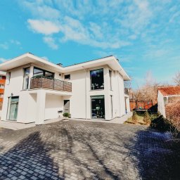 MONTOWNIA DOMÓW SPÓŁKA Z OGRANICZONĄ ODPOWIEDZIALNOŚCIĄ - Nowoczesny, dwukondygnacyjny dom z płaskim dachem, białymi ścianami i dużymi oknami, widok z zewnątrz na podjazd z kostki brukowej w cieniu drzew, błękitne niebo z chmurami w tle.