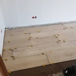 MONTOWNIA DOMÓW SPÓŁKA Z OGRANICZONĄ ODPOWIEDZIALNOŚCIĄ - Podłoga w trakcie montażu paneli z użyciem brązowego kleju i żółtych krzyżyków dystansowych, widoczne gniazdka elektryczne na białej ścianie.