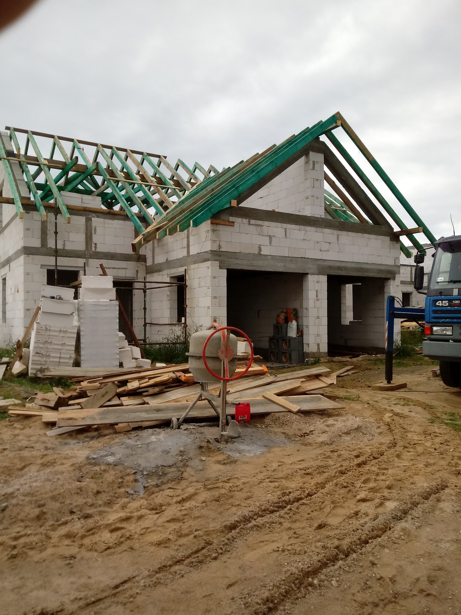 Budowa domu jednorodzinnego w trakcie wznoszenia konstrukcji dachu, widoczna betoniarka, bloczki Ytong, drewniane elementy konstrukcyjne i ciężarówka z podnośnikiem.