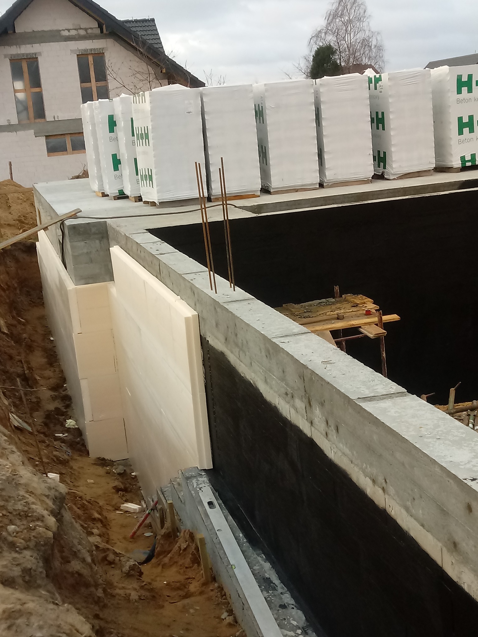 Budowa fundamentów: ściana fundamentowa z izolacją termiczną, zabezpieczona hydroizolacją, z widocznymi zbrojeniami i paletami bloczków betonowych H+H na górze.