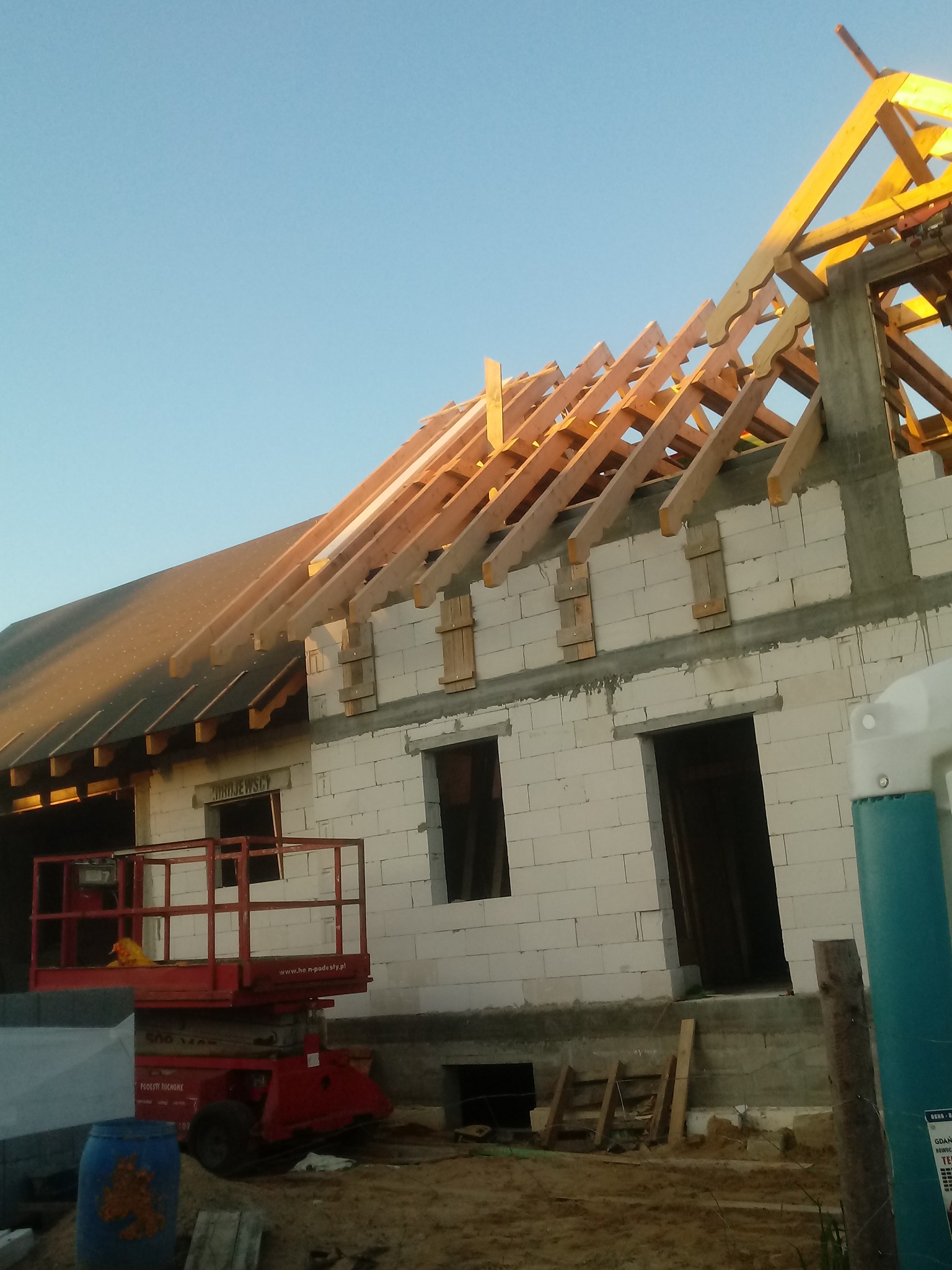 Surowy stan domu z białego betonu komórkowego w trakcie budowy dachu z drewnianą konstrukcją więźby, widoczny podnośnik koszowy i częściowo wykonane deskowanie.