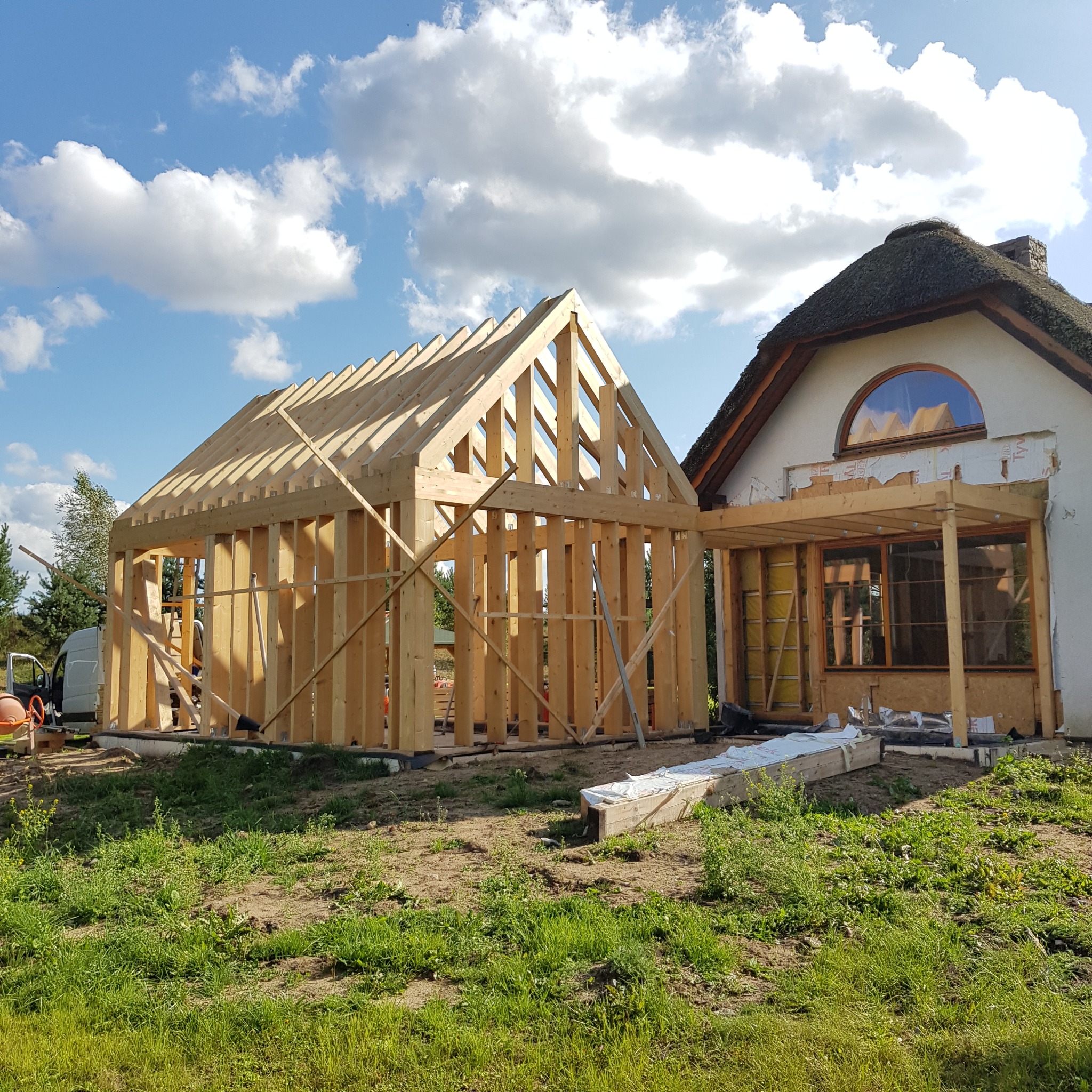 Surowa konstrukcja drewniana dobudowy domu z dachem krytym strzechą w słoneczny dzień, widoczne materiały budowlane i betoniarka.