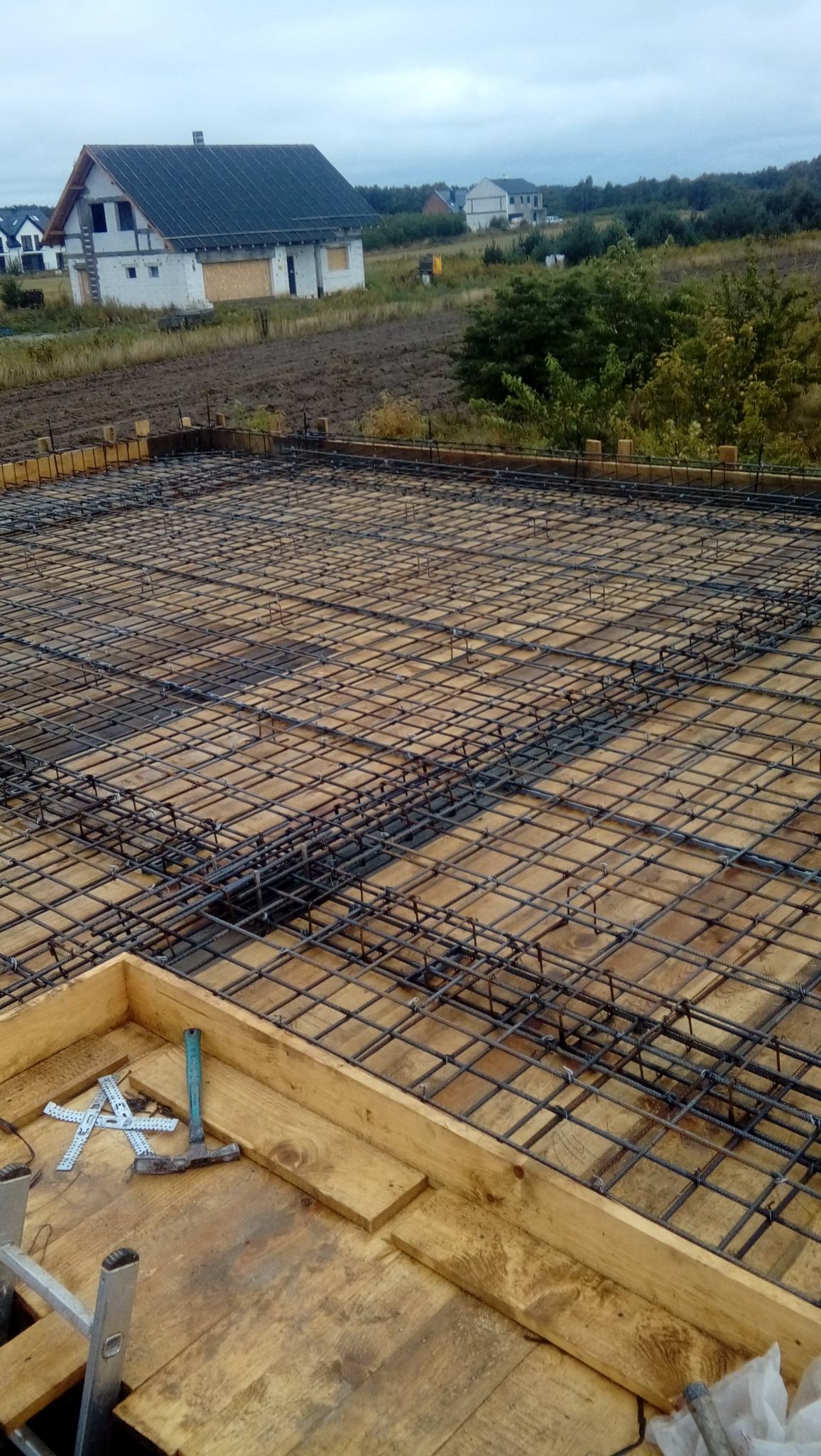 Zbrojenie płyty fundamentowej z deskowaniem i narzędziami w rogu, w tle budowa domu jednorodzinnego w stanie surowym otwartym.