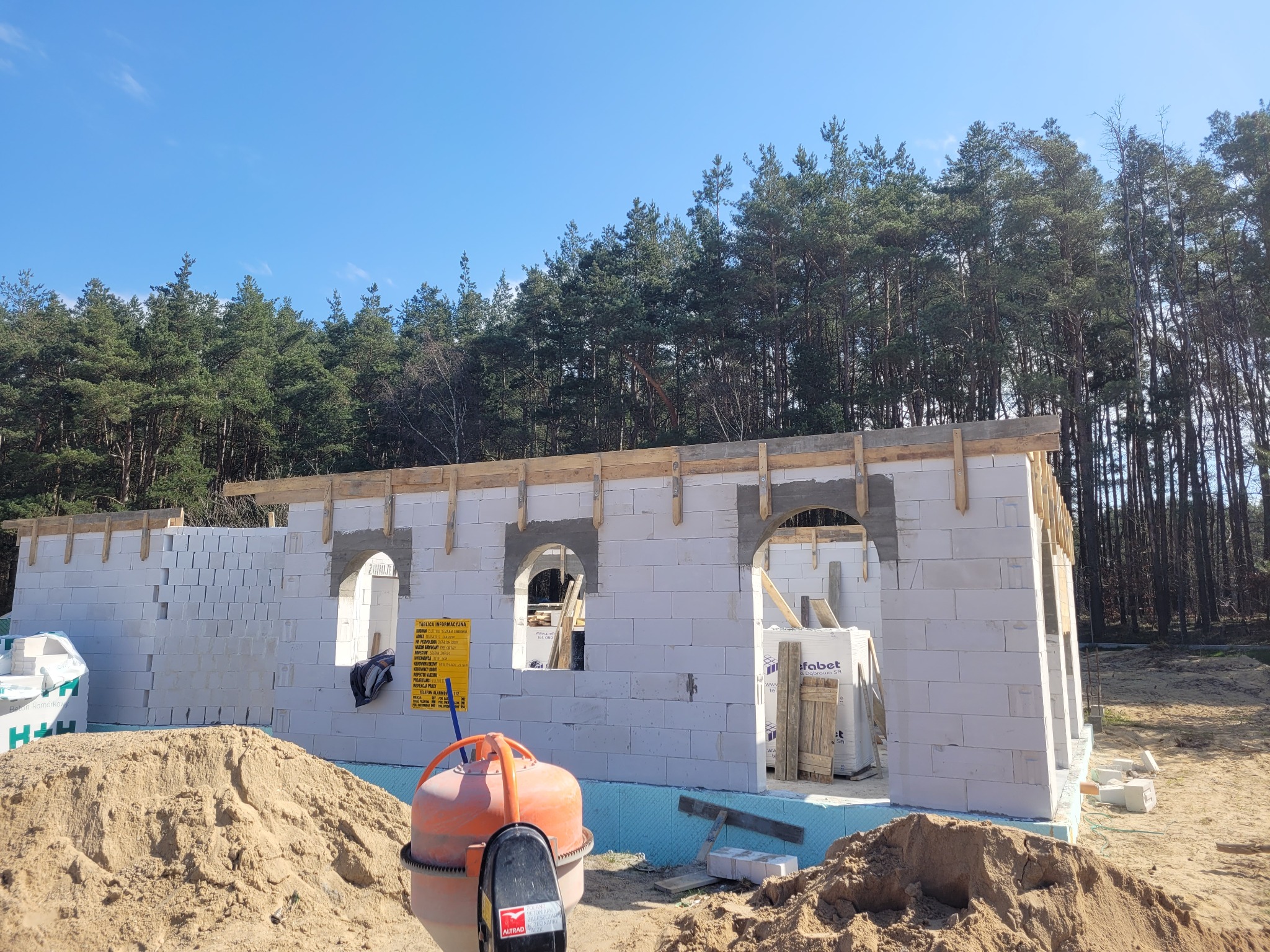 Wznoszenie ścian parteru domu murowanego z bloczków betonu komórkowego, widoczne nadproża okienne w kształcie łuków, mieszarka do betonu na pierwszym planie, w tle las iglasty i błękitne niebo.