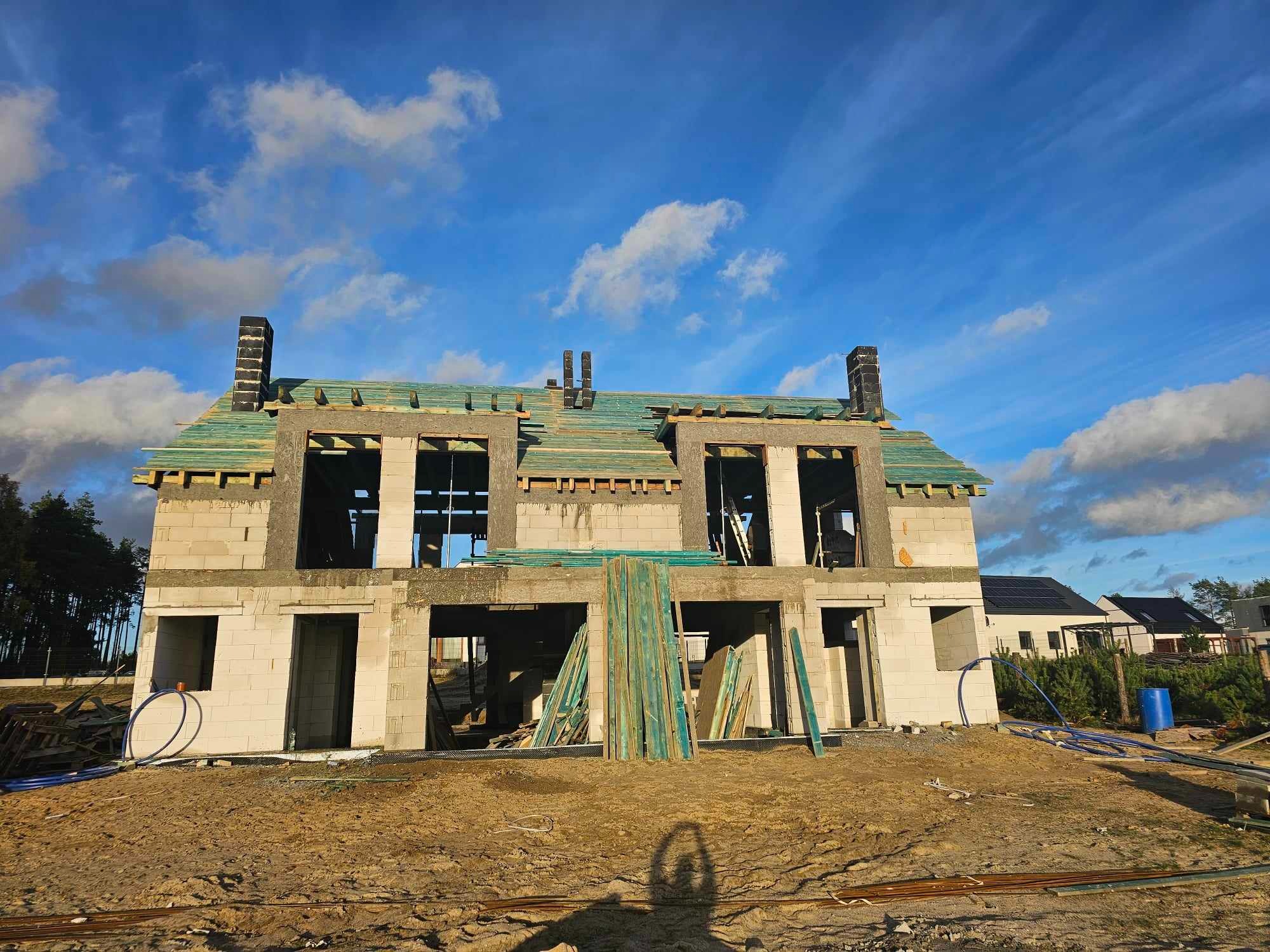 Dwukondygnacyjny budynek w stanie surowym otwartym z widocznym drewnianym szkieletem dachu, kominami z cegły klinkierowej i materiałami budowlanymi na placu budowy w słoneczny dzień.