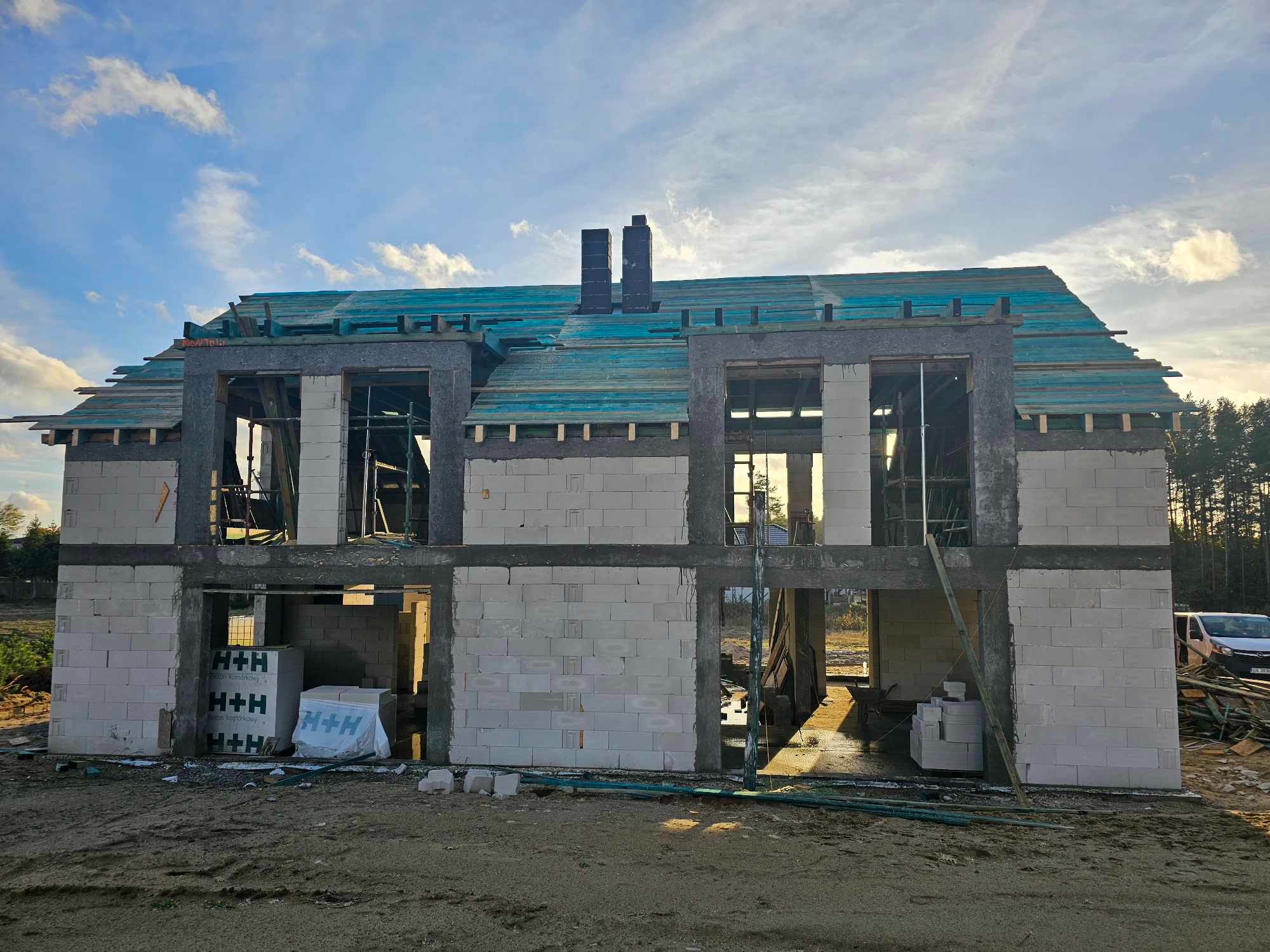 Dwukondygnacyjny dom w trakcie budowy, wzniesiony z białych bloczków H+H, z widocznym deskowaniem dachu pokrytym turkusową folią, na tle błękitnego nieba z chmurami i lasu.