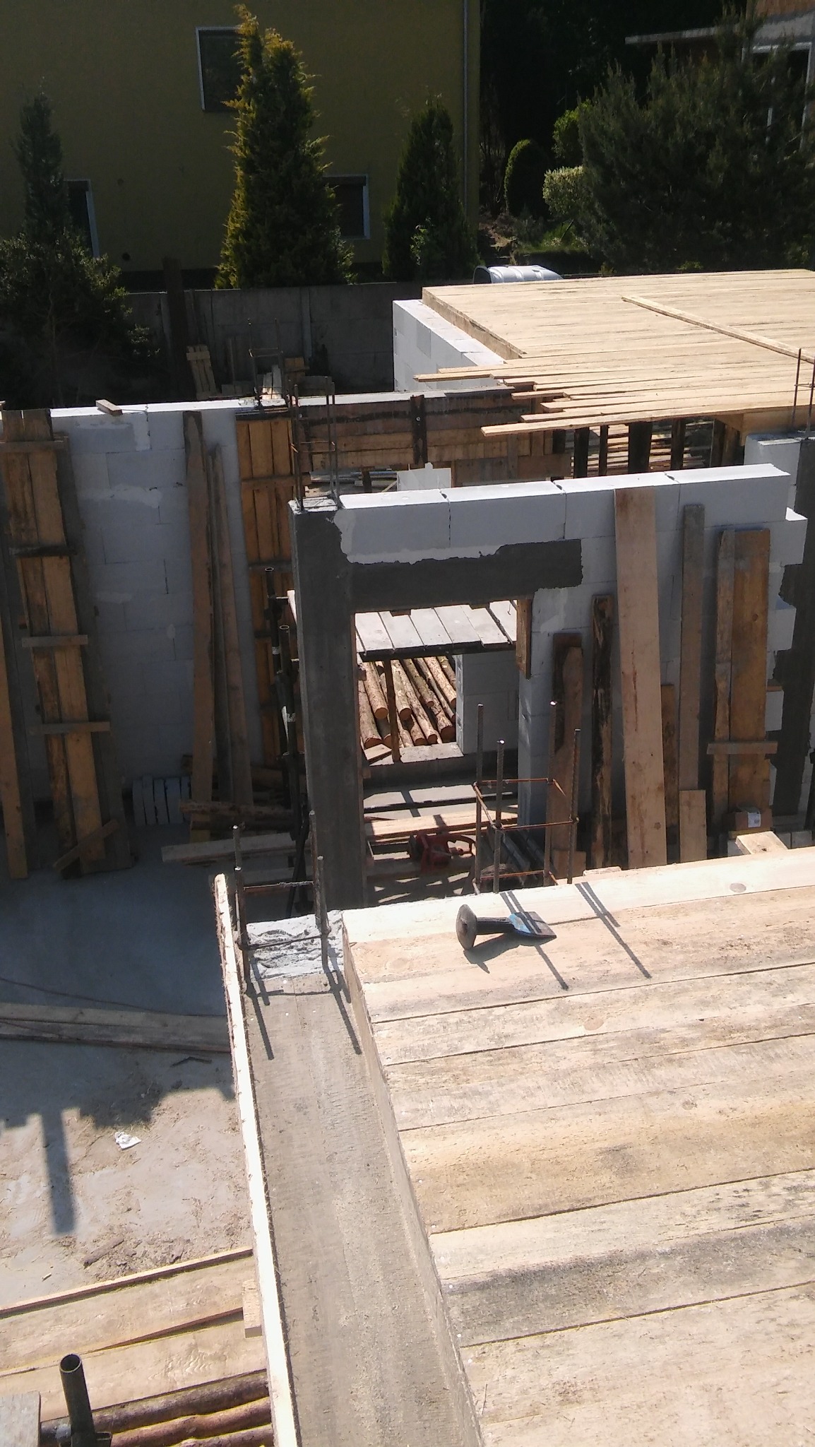 Wznoszenie ścian domu z bloczków betonowych, widoczne zbrojenie i szalunki, częściowo deskowane, w tle zielona trawa i drzewa.