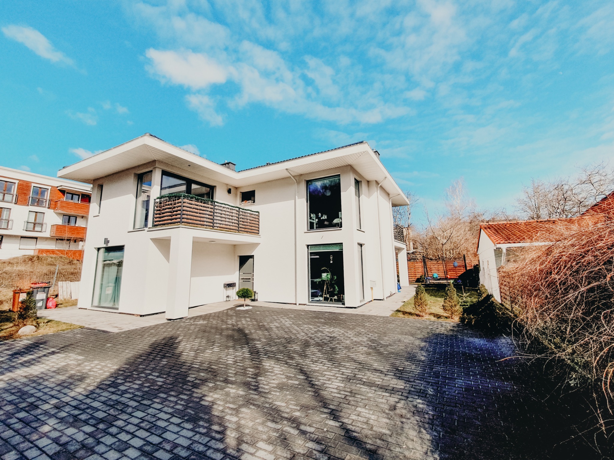 Nowoczesny, dwukondygnacyjny dom z płaskim dachem, białymi ścianami i dużymi oknami, widok z zewnątrz na podjazd z kostki brukowej w cieniu drzew, błękitne niebo z chmurami w tle.