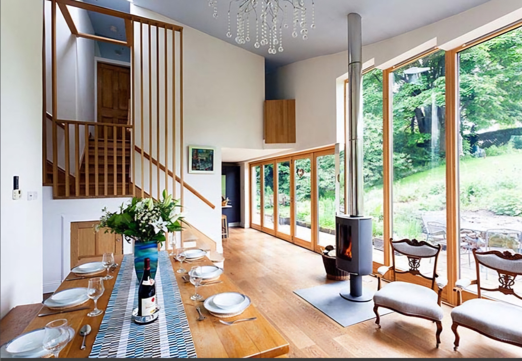 Jasne, przestronne wnętrze domu z drewnianą podłogą, dużymi oknami z widokiem na ogród, nowoczesnym kominkiem, drewnianymi schodami i stołem nakrytym do posiłku.