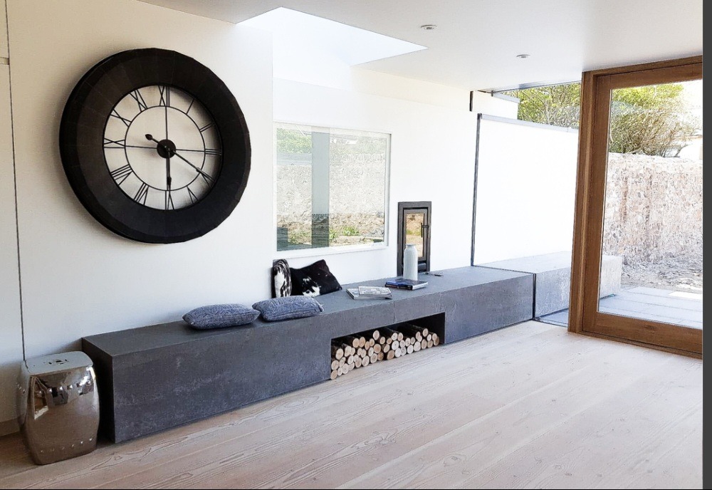 Jasne wnętrze z drewnianą podłogą, szarą zabudową z miejscem na drewno kominkowe, dużym zegarem na ścianie i wyjściem na taras.