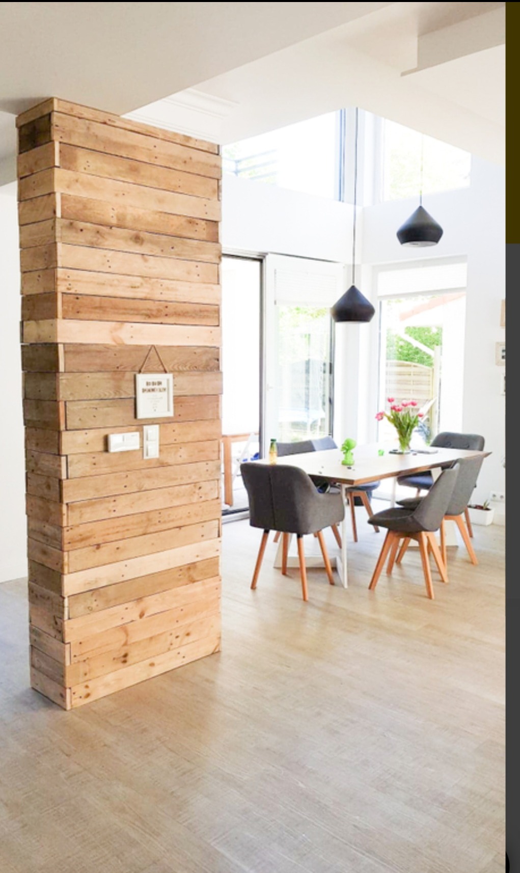 Drewniana kolumna z palet oddzielająca przestrzeń jadalną od reszty pomieszczenia, widoczna jasna podłoga i stół z krzesłami w tle, lampy wiszące.