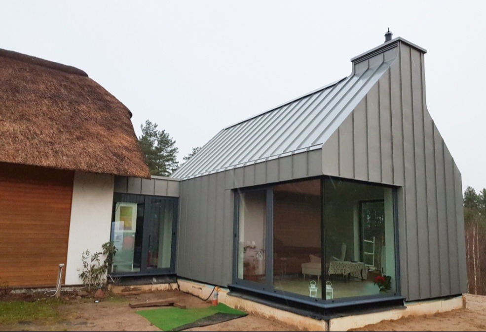 Nowoczesny dom z dachem krytym blachą na rąbek stojący, połączony z budynkiem o tradycyjnym strzechą, widok z zewnątrz w pochmurny dzień.