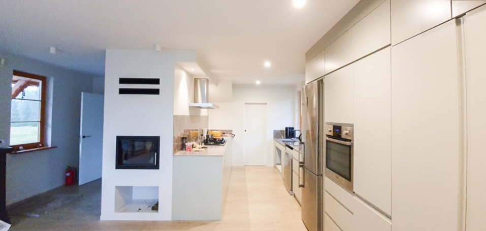 Wnętrze domu w trakcie wykańczania: widoczny kominek z czarnym wkładem, jasna kuchnia z zabudową meblową, okno z drewnianą ramą i widok na zewnątrz, częściowo wykończona podłoga i czerwona gaśnica...