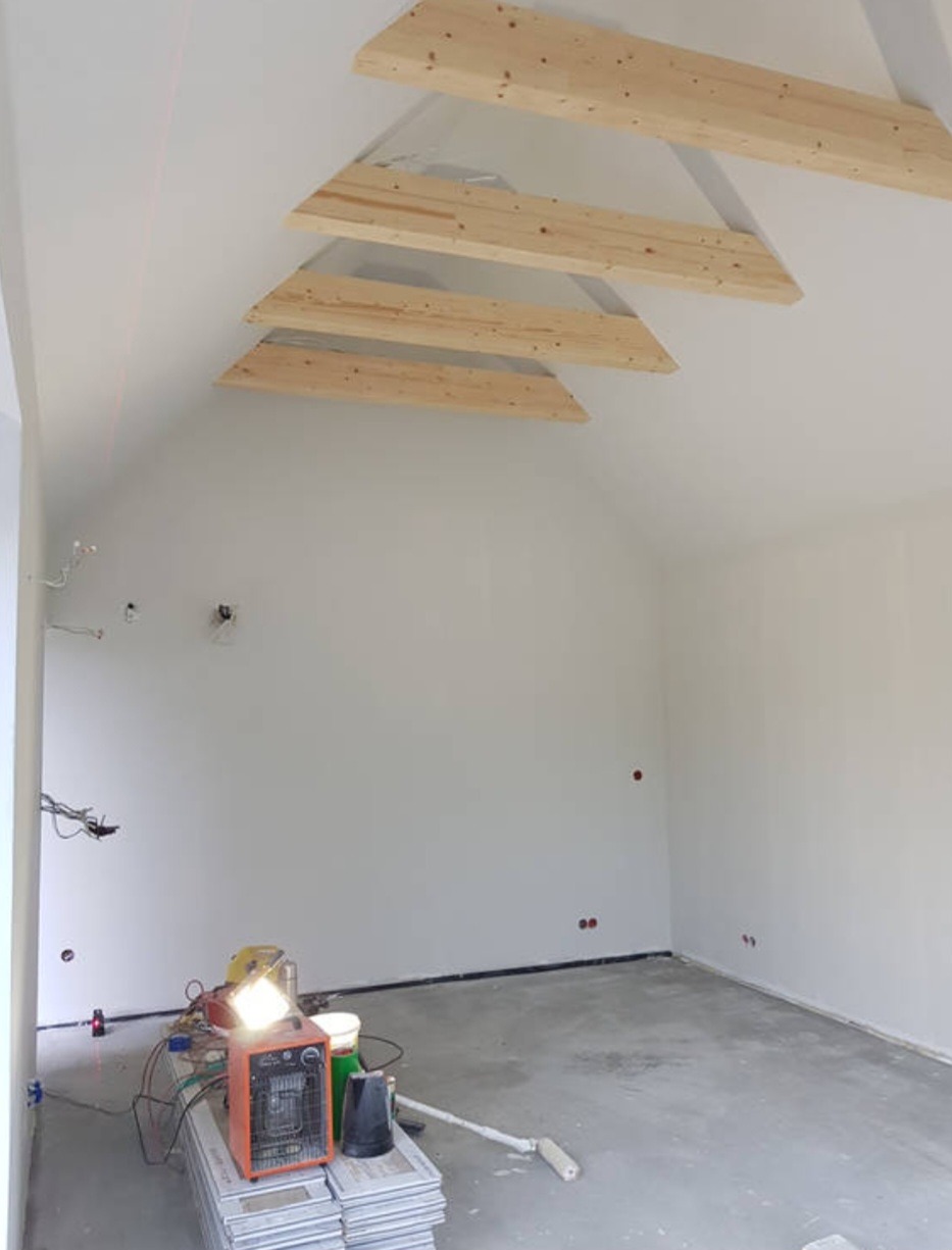 Surowe wnętrze poddasza z widocznymi drewnianymi elementami konstrukcyjnymi dachu, świeżo pomalowanymi ścianami i betonową podłogą, z narzędziami i grzejnikiem budowlanym w rogu.
