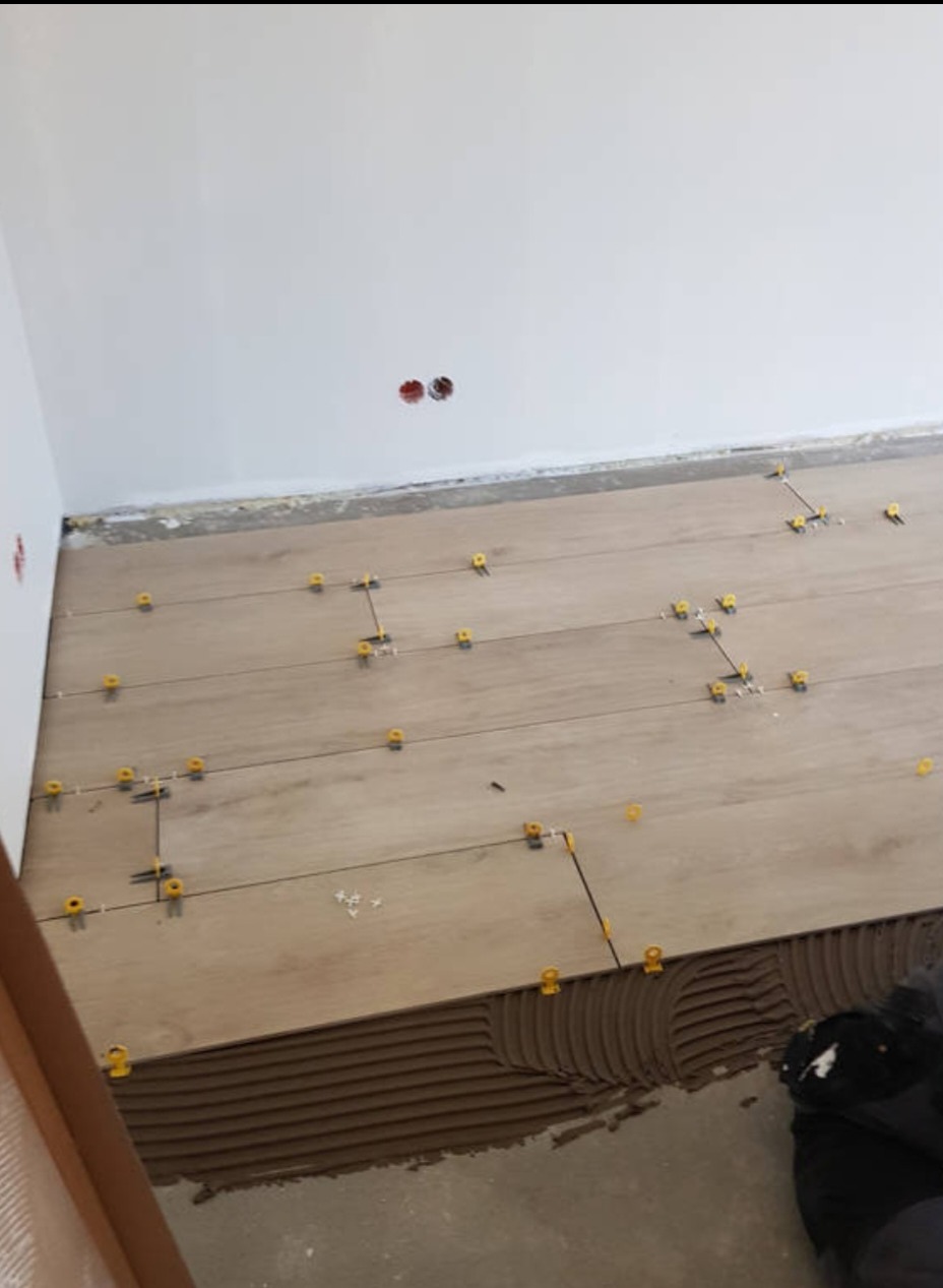 Podłoga w trakcie montażu paneli z użyciem brązowego kleju i żółtych krzyżyków dystansowych, widoczne gniazdka elektryczne na białej ścianie.