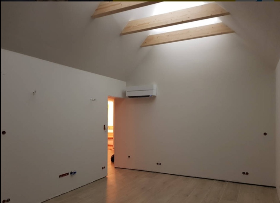 Poddasze po remoncie: świeżo pomalowane ściany w odcieniach bieli, drewniane belki stropowe, klimatyzator na ścianie, widoczne punkty elektryczne i jasna podłoga z paneli.