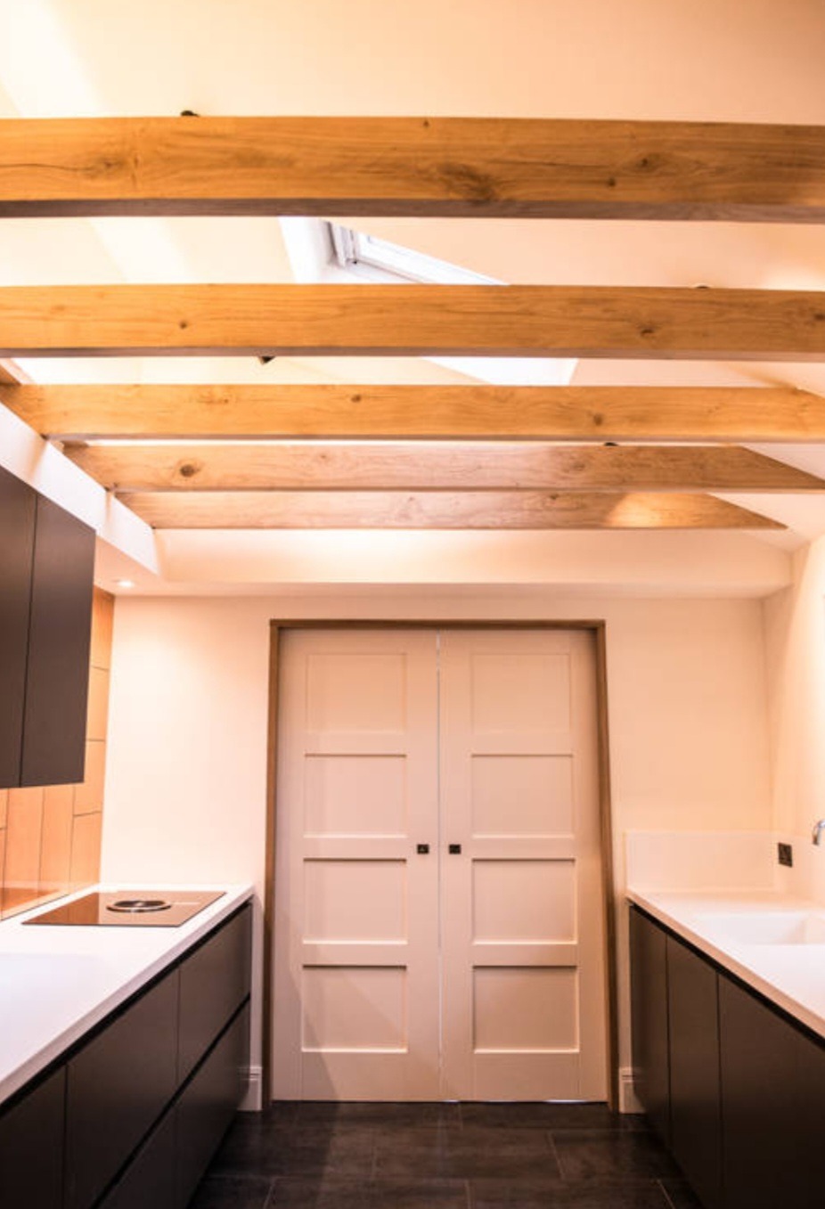 Nowoczesna kuchnia z drewnianymi belkami stropowymi, minimalistycznymi szafkami w kolorze grafitu, białym blatem i szarymi płytkami podłogowymi, widziana z perspektywy podłogi.