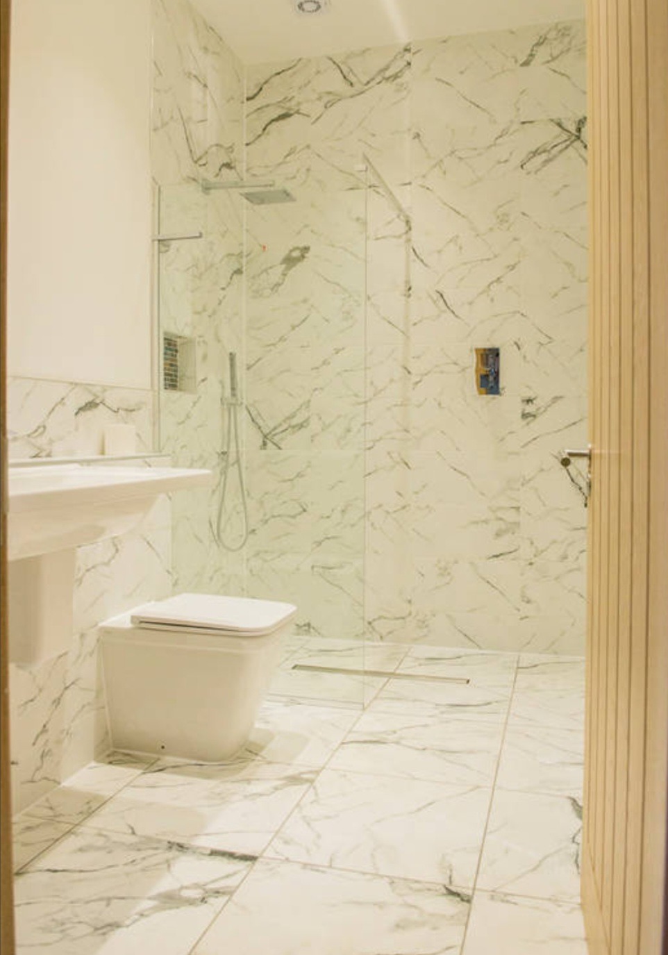 Nowoczesna łazienka z białymi płytkami imitującymi marmur na ścianach i podłodze, kabina prysznicowa typu walk-in z deszczownicą, umywalka nablatowa i wisząca toaleta, widok zza drewnianych drzwi.