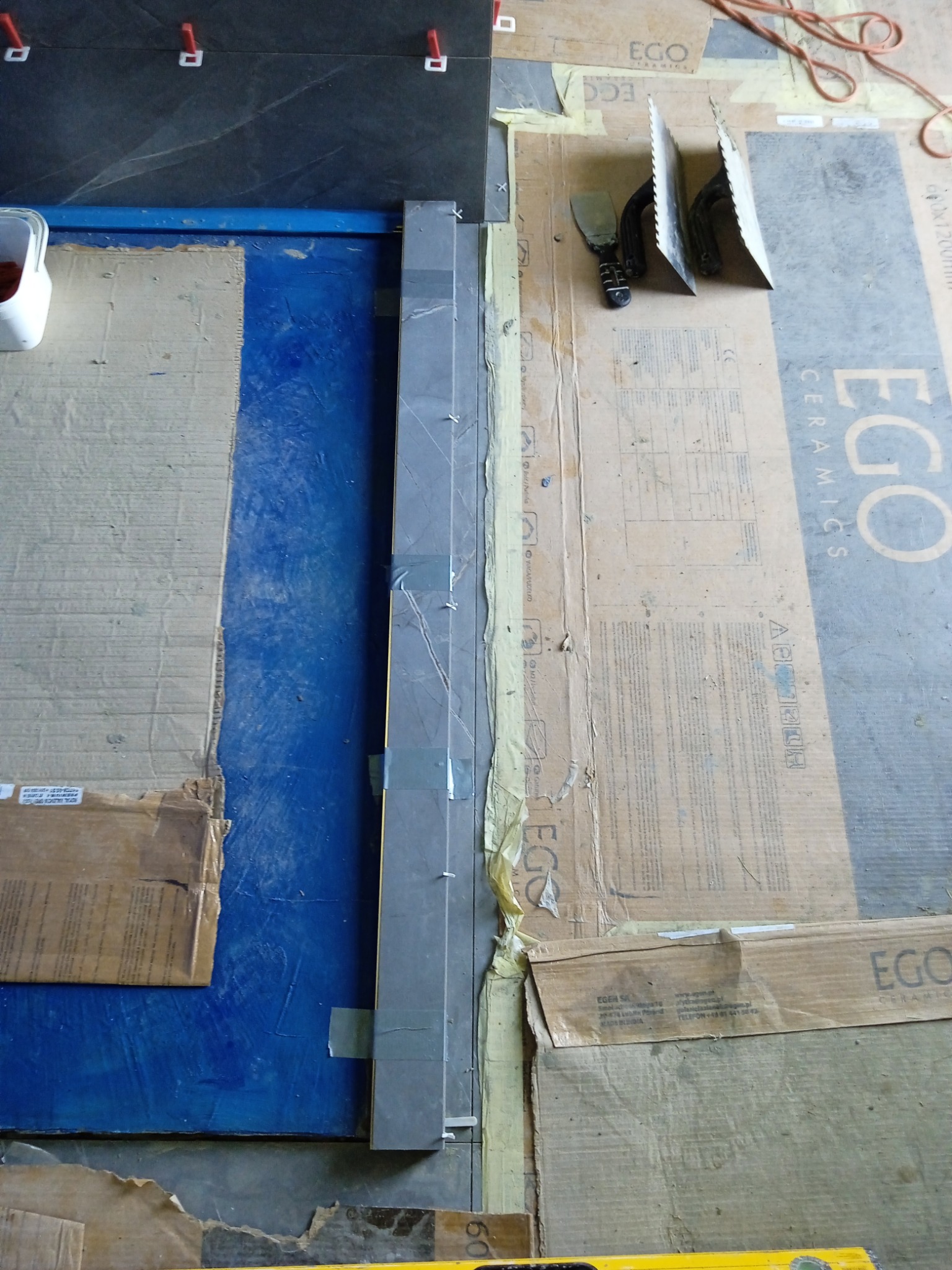 Układanie ciemnoszarych płytek na podłodze, widoczne narzędzia glazurnicze, poziomica, kartony zabezpieczające podłoże i krzyżyki dystansowe.