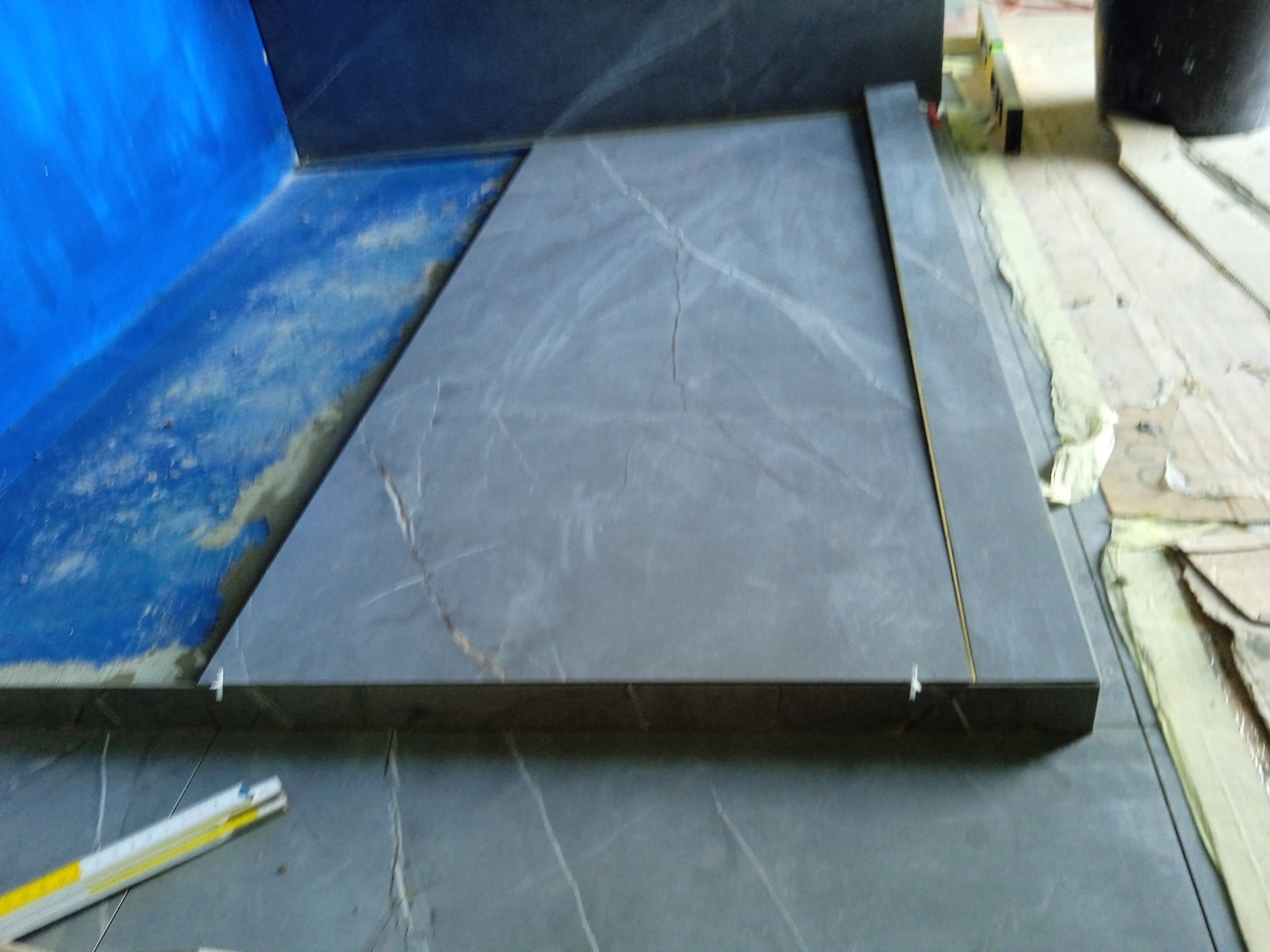 Układanie dużej, szarej płytki imitującej marmur w trakcie prac wykończeniowych, widoczne plastikowe krzyżyki dystansowe i poziomica.