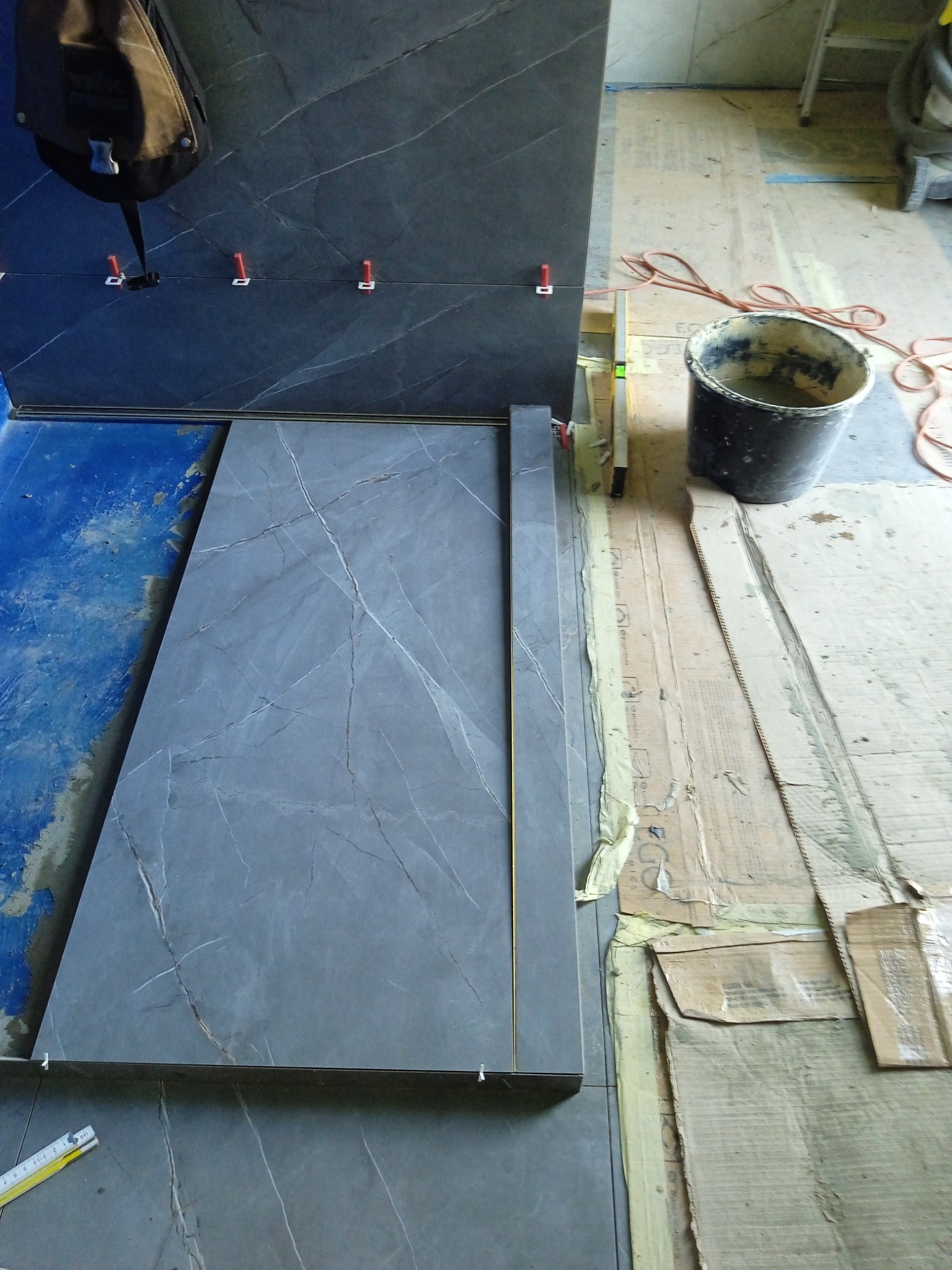 Układanie wielkoformatowych, grafitowych płytek imitujących marmur z użyciem poziomicy i klipsów dystansowych; widoczny wiadro z zaprawą i torba robocza.