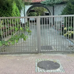 Dwuskrzydłowa brama wjazdowa z metalowymi prętami, szara, z zielenią w tle i kostką brukową na podjeździe, widok z zewnątrz.