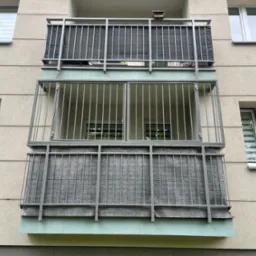 Dwa balkony z metalowymi balustradami i szarą osłoną, zamontowane na elewacji budynku z oknami. Górny balkon z budką dla ptaków.