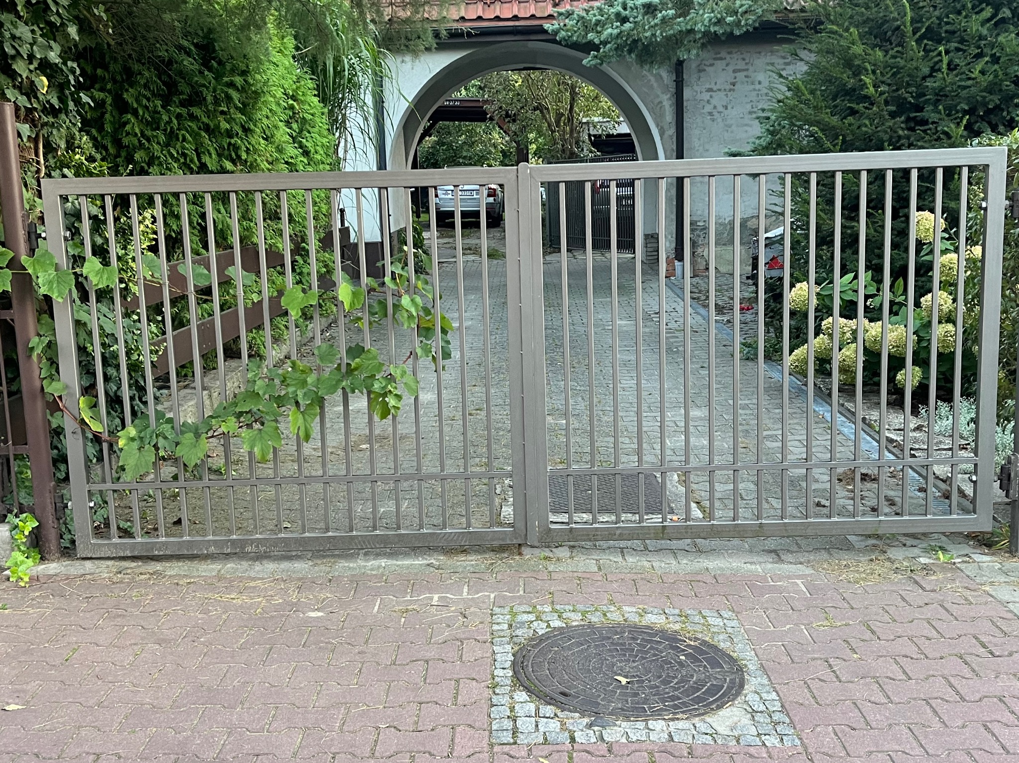 Dwuskrzydłowa brama wjazdowa z metalowymi prętami, szara, z zielenią w tle i kostką brukową na podjeździe, widok z zewnątrz.